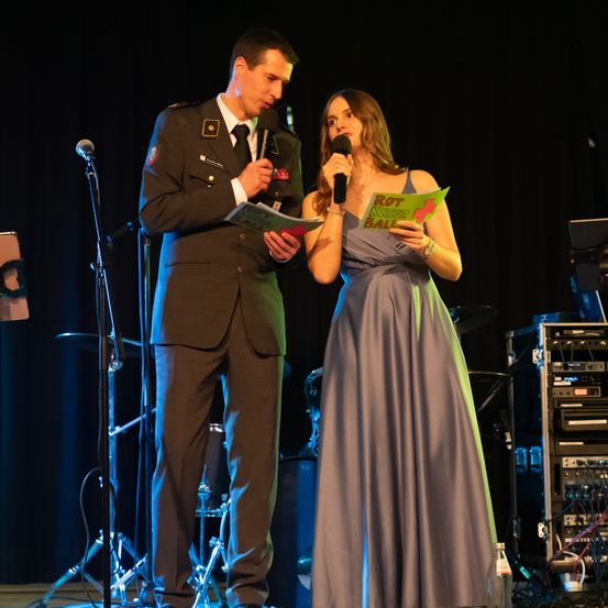 Ein Mann in Uniform und eine Frau in einem Kleid stehen auf der Bühne, halten Bücher und Mikrofone. Der Mann trägt eine Krawatte und die Frau ein Armband. Sie stehen auf einer Bühne mit schwarzem Hintergrund.