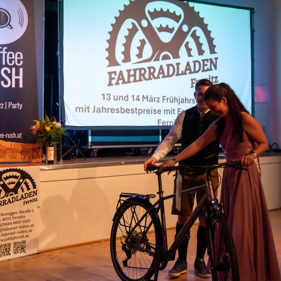 Zwei Personen stehen auf der Bühne und präsentieren ein Fahrrad. Eine Frau hält das Lenker und ein Mann steht hinter ihr. Eine Projektionsleinwand im Hintergrund zeigt 'Fahrradladen' und Termine für die Veranstaltung.