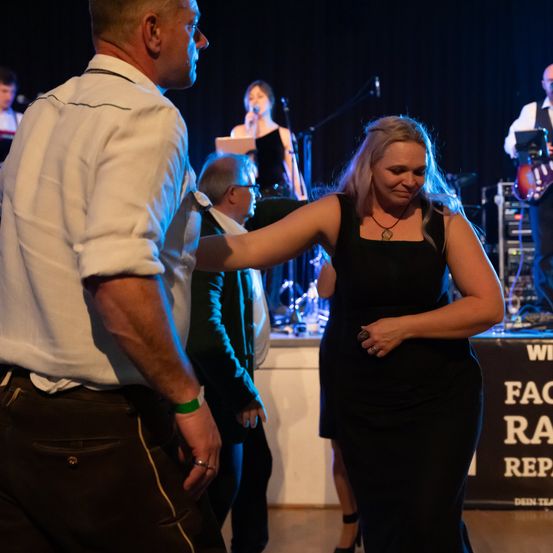 Eine Frau in einem schwarzen Kleid tanzt mit einem Mann in einem weißen Hemd und schwarzen Hosen bei einem Konzert. Eine Band spielt im Hintergrund.