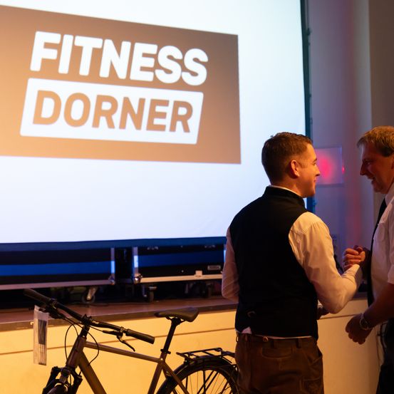 Zwei Männer stehen vor einer großen Leinwand, auf der 'Fitness Dorner' zu sehen ist. Der Mann auf der linken Seite schüttelt dem anderen Mann die Hand. Ein Fahrrad ist in der Nähe geparkt.