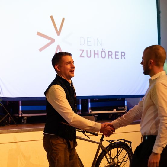 Zwei Männer schütteln sich die Hände vor einer Bühne mit einer Projektionsleinwand im Hintergrund, auf der der Text 'Dein Zuhörer' zu sehen ist. Der Mann auf der linken Seite lächelt.