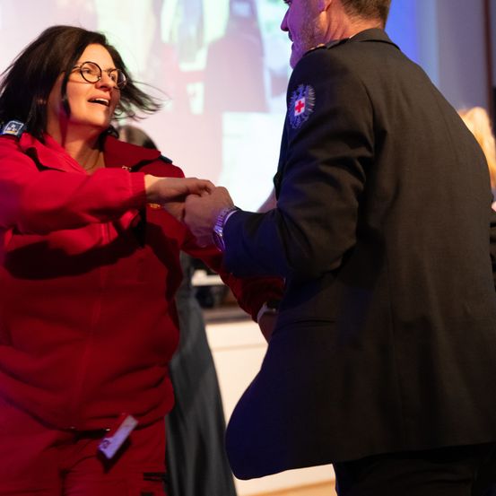 Eine Frau in einer roten Uniform schüttelt die Hand eines Mannes in einer Marineuniform mit einem roten Kreuz-Patch auf seiner Schulter.