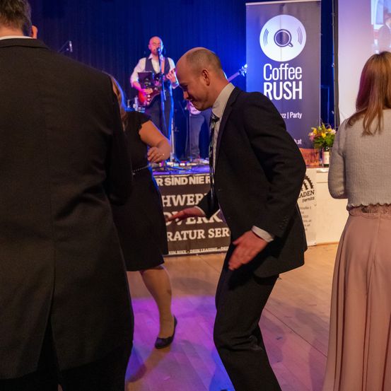 Ein Mann in einem Anzug tanzt auf der Bühne bei einer Coffee Rush-Veranstaltung. Ein Gitarrist und ein Sänger spielen im Hintergrund. Ein anderer Mann und eine Frau stehen im Vordergrund.
