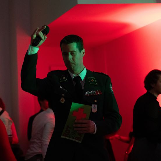 Ein Mann in Uniform spricht in ein Mikrofon und hält ein Büchlein in der Hand, mit roter Beleuchtung im Hintergrund.