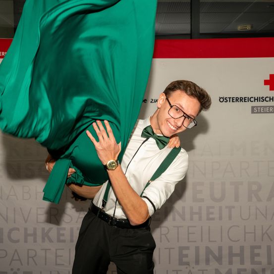 Ein Mann in einem weißen Hemd mit grünen Trägern hält eine Frau in einem grünen Kleid mit beiden Händen. Die Frau hält den Kopf des Mannes. Sie lächeln beide.