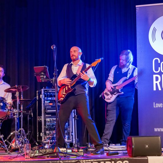 Eine Band spielt auf der Bühne vor einem blauen Vorhang. Ein Gitarrist spielt eine E-Gitarre, während ein Bassist eine Bassgitarre spielt. Ein Schlagzeuger steht hinter einem Schlagzeug. Ein Mikrofon steht vor dem Gitarristen auf einem Ständer.