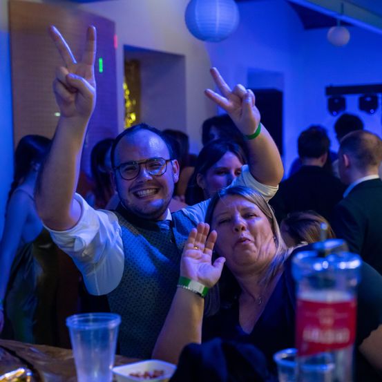 Eine Gruppe von Menschen auf einer Party, ein Mann und eine Frau machen ein Friedenszeichen, lächeln und posieren für ein Foto.