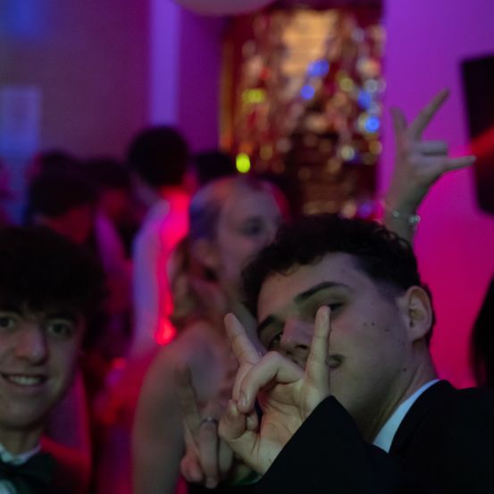 Ein Mann posiert mit seinen Fingern in einem Friedenszeichen auf einer lebhaften Party, umgeben von anderen Gästen unter einem leuchtenden rosa Licht.