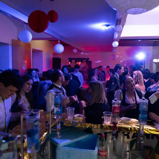 Eine Party mit Leuten in einem Raum mit blauen und roten Lichtern, Flaschen und Gläsern auf einem Tisch. Es hängen Papierlaternen an der Decke.