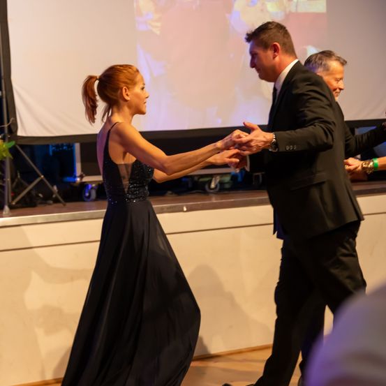 Eine Frau in einem schwarzen Kleid tanzt mit einem Mann in einem Anzug. Sie lächeln beide und halten sich an den Händen. Dahinter ist eine große Leinwand.