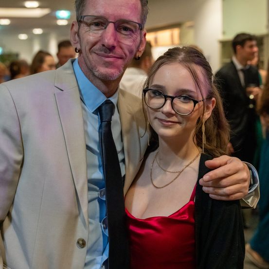 Ein Mann und eine Frau lächeln für ein Foto bei einer Veranstaltung. Der Mann trägt einen Anzug und eine Krawatte, und die Frau trägt ein rotes Kleid und eine Brille.