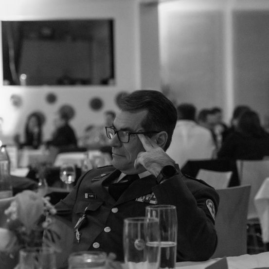 Ein Mann in Uniform sitzt an einem Tisch, mit einer Hand am Kopf und Gläsern auf dem Tisch. Es gibt leere Gläser und Flaschen auf dem Tisch. Im Hintergrund sitzen Menschen an anderen Tischen.