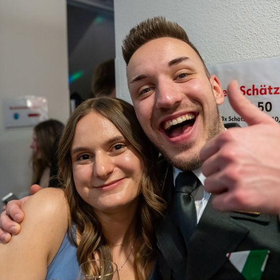 Ein Mann und eine Frau lächeln und posieren für ein Foto mit Partyhintergrund. Der Mann zeigt den Daumen nach oben, während die Frau ein Glas hält.
