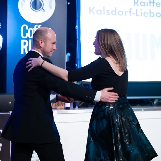 Ein Mann und eine Frau tanzen zusammen auf einer Bühne vor einer Leinwand mit einem Kaffeelogo.