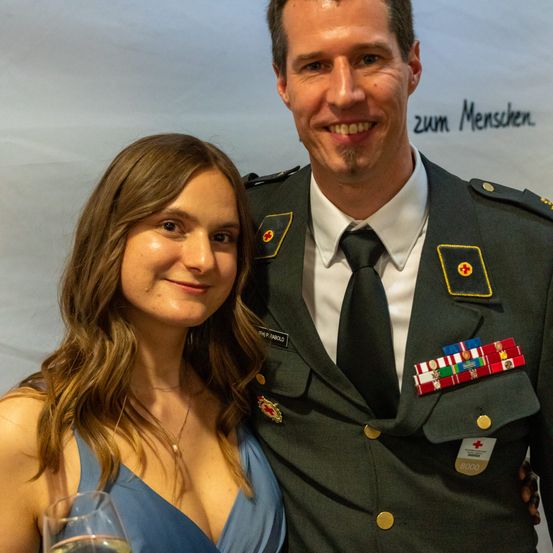 Ein Mann in Uniform und eine Frau in einem blauen Kleid lächeln und stehen eng beieinander.