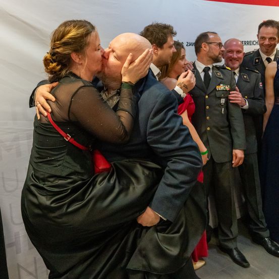 Ein Paar küsst sich, umgeben von anderen, einige in Uniformen. Ein weißer Hintergrund steht hinter ihnen.