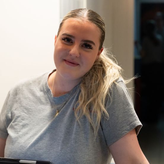 Eine Frau mit blonden Haaren und einem grauen T-Shirt lächelt und posiert für ein Foto. Sie trägt eine Halskette und sitzt vor einer weißen Wand.