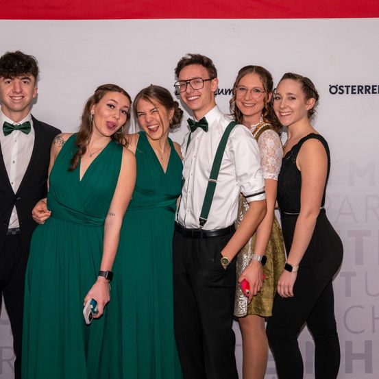 Fünf Personen, feierlich gekleidet, stehen eng beieinander für ein Foto und lächeln. Drei Frauen tragen grüne Kleider, und zwei Männer tragen formelle Anzüge. Der Hintergrund hat einen weißen Hintergrund mit Text.