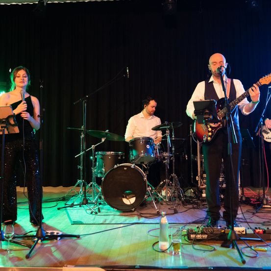 Eine Band tritt auf der Bühne auf, bestehend aus einem Schlagzeuger, einem Gitarristen und einem Sänger. Die Frau steht links, der Schlagzeuger in der Mitte und der Gitarrist rechts.