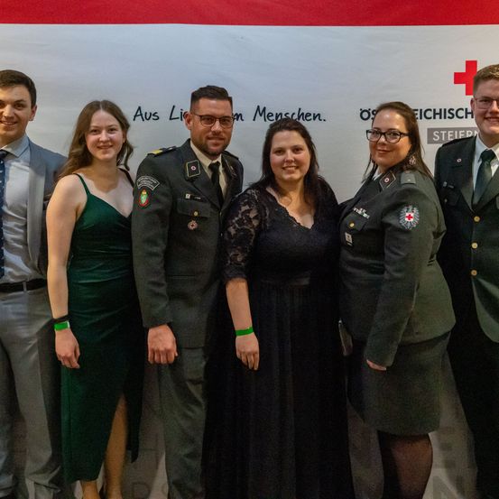 Eine Gruppe von Menschen, darunter Militärangehörige und Zivilisten, steht für ein Foto zusammen, mit einem Banner im Hintergrund. Sie alle scheinen zu lächeln.