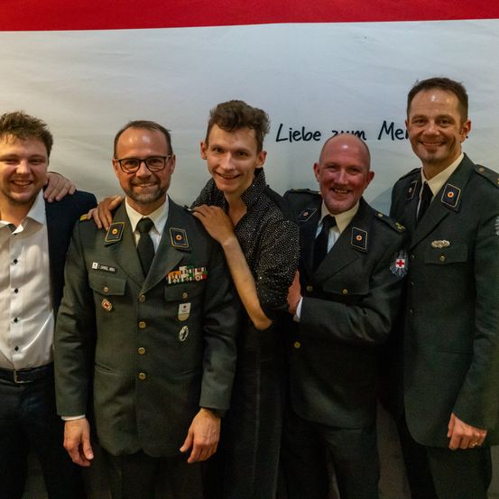 Fünf Männer stehen vor einem weißen Banner mit deutschem Text. Sie lächeln alle und scheinen für ein Foto zu posieren.