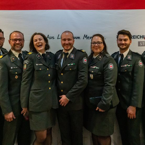 Sechs Personen in Militäruniformen lächeln und posieren für ein Foto vor einem weißen Banner mit rotem Text.
