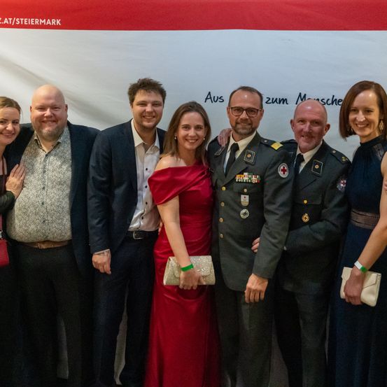Eine Gruppe von sieben Personen posiert für ein Foto vor einem Banner mit Text. Die Frau in einem roten Kleid hält eine Handtasche. Die Männer tragen Anzüge und Militäruniformen.