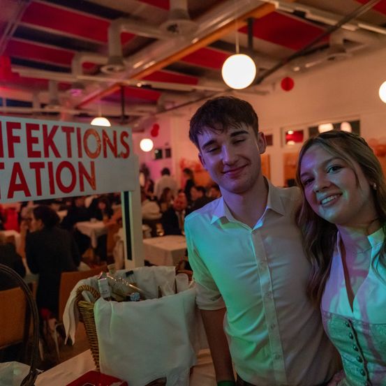 Ein Paar steht neben einem Schild mit der Aufschrift 'Infektionsstation', möglicherweise auf einer Party, mit bunten Lichtern und einer festlichen Atmosphäre. Im Hintergrund sitzen Leute an Tischen.