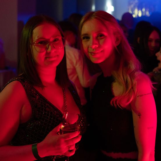 Zwei Frauen stehen in einem Club mit roter Beleuchtung. Die Frau auf der linken Seite trägt eine Brille und hält ein Weinglas. Die Frau auf der rechten Seite hat eine Tätowierung am Arm.