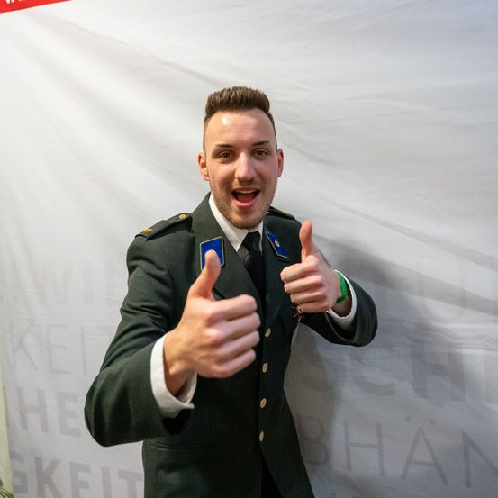 Ein lächelnder Mann in Uniform gibt ein Daumen-hoch-Zeichen. Im Hintergrund steht ein weißer Vorhang mit den Worten 'Freiheit' und 'Sicherheit'.