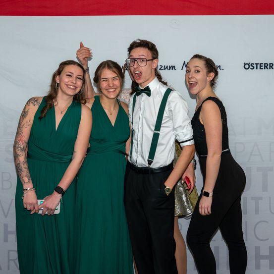 Vier Personen stehen zusammen auf einem roten Teppich und lächeln. Zwei Frauen tragen grüne Kleider, während ein Mann in einem weißen Hemd und suspenders zwischen ihnen steht. Eine Frau in Schwarz hält ein Handy. Sie posieren für ein Foto vor einem Banner.
