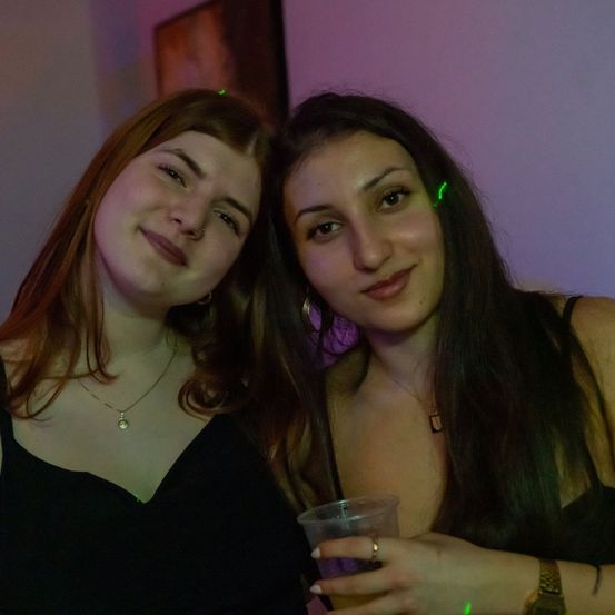 Zwei Frauen posieren für ein Foto auf einer Party. Beide tragen schwarze Kleider und halten Getränke. Eine hat eine Halskette mit einem Anhänger, und die andere trägt eine Uhr. Eine Wand mit einem Bilderrahmen ist hinter ihnen.