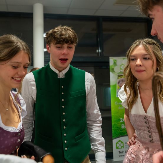 Drei junge Menschen stehen in einem Raum beieinander. Ein Mann in einer grünen Weste steht zwischen zwei Frauen. Eine Frau trägt eine Halskette und die andere ein weißes Kleid. Sie alle lächeln. Im Hintergrund befindet sich ein Poster an der Wand.