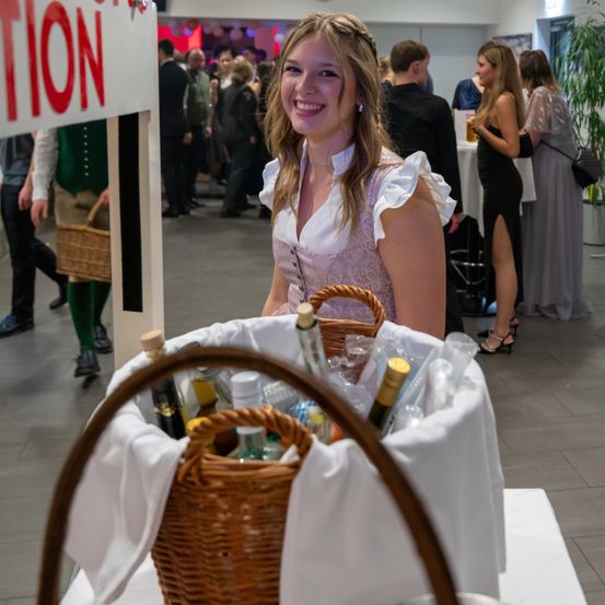Eine lächelnde Frau in traditioneller Kleidung steht neben einem Korb mit Flaschen bei einer Veranstaltung, mit einem Schild, das 'TION' dahinter steht. Andere Gäste sind im Hintergrund zu sehen.