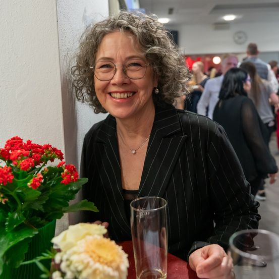 Eine lächelnde Frau mit lockigem Haar und Brille sitzt an einem Tisch mit Blumen, Gläsern und einem geschäftigen Hintergrund.
