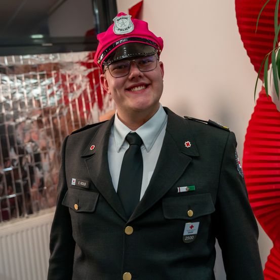 Eine lächelnde Person in einem schwarzen Uniform mit einem roten Hut, einer Brille und einem Namensschild mit dem Namen E. Kolb.