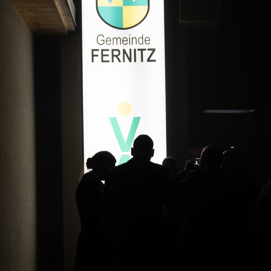 Eine Gruppe von Menschen steht vor einem großen, teilweise beleuchteten Schild, auf dem das Wort 'Gemeindel' sichtbar ist.