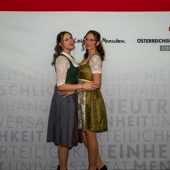Zwei Frauen in traditioneller Kleidung, eine in Grün und die andere in Grau, stehen eng beieinander vor einem Banner mit der Aufschrift 'Österreichische'. Sie lächeln und posieren für ein Foto.