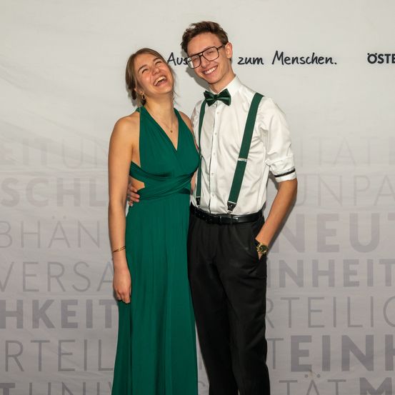 Ein junger Mann und eine junge Frau stehen lächelnd eng beieinander. Die Frau trägt ein grünes Kleid und der Mann ein weißes Hemd mit grünen Hosenträger und eine Fliege. Hinter ihnen zeigt ein Hintergrund Text in einer fremden Sprache.