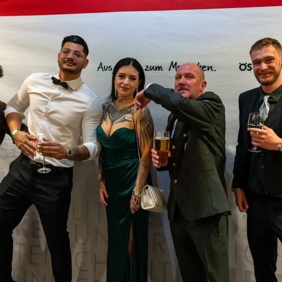 Fünf Personen stehen bei einer Veranstaltung eng beieinander. Der Mann in der Mitte hält ein Glas Bier. Die Frau links von ihm trägt ein grünes Kleid und der Mann rechts von ihm hält ein Glas Wein. Der Hintergrund ist ein Banner mit schwarzem Text.