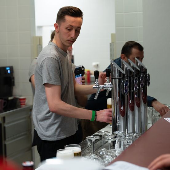 Ein Mann in einem grauen T-Shirt arbeitet an einer Bar, hält einen Zapfhahn und füllt Bier in Gläser. Ein anderer Mann beobachtet ihn, und verschiedene Gläser und Bierzapfhähne sind auf der Theke angeordnet.