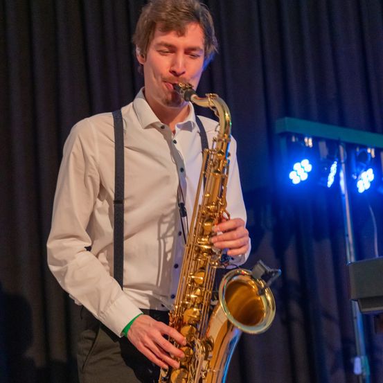 Ein Mann spielt Saxophon auf der Bühne, beleuchtet von blauen Lichtern, mit einem schwarzen Vorhang im Hintergrund.