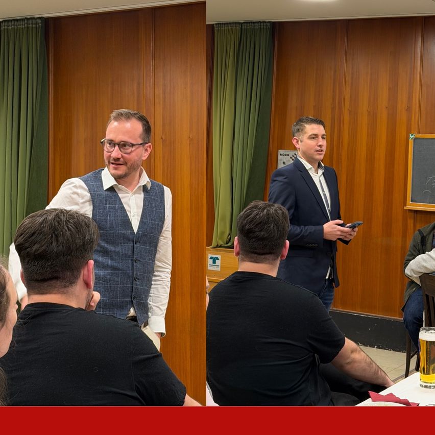 Zwei Männer sprechen miteinander. Ein Mann steht und hält ein Telefon. Ein Mann sitzt mit einem Bier vor sich.