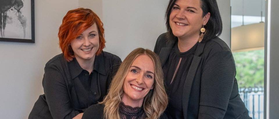 Drei Frauen in schwarzen Outfits posieren in einem Büro. Die Frau in der Mitte lächelt und sitzt auf einem Stuhl, während die anderen hinter ihr stehen. Ein Bilderrahmen hängt an der Wand.