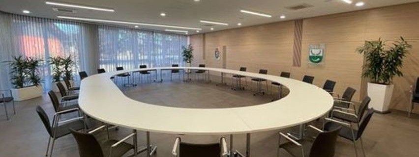 Ein großer Konferenzraum verfügt über einen langen, gebogenen Tisch, der von schwarzen Stühlen umgeben ist. Holzwände und große Fenster mit Jalousien sind vorhanden. Zwei Topfpflanzen befinden sich auf beiden Seiten des Raumes.