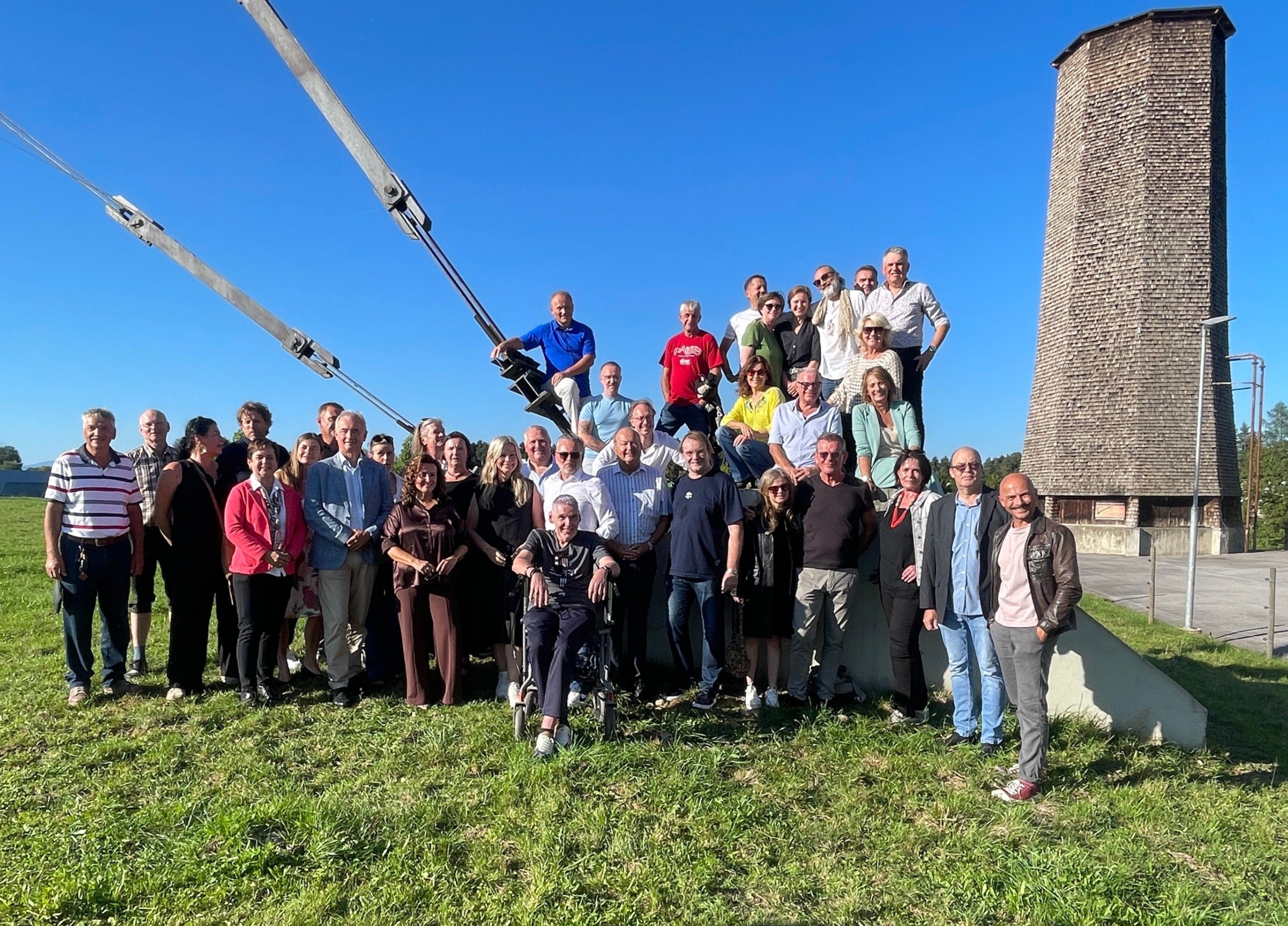 Eine Gruppe von Menschen steht im Freien auf einem Grasfeld und posiert für ein Foto. Ein Mann im Rollstuhl ist zentral positioniert. Einige tragen Sonnenbrillen und einige tragen Hüte. Im Hintergrund befindet sich eine große Struktur.
