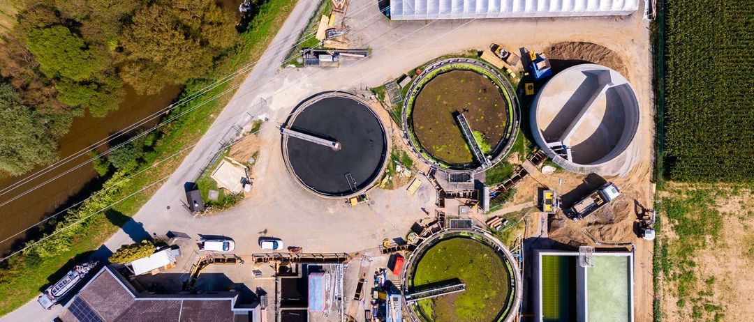 Luftaufnahme einer Wasseraufbereitungsanlage. Sie verfügt über mehrere große Tanks, einer mit grüner Flüssigkeit. In der Nähe befinden sich Solarmodule auf einem Gebäude. Das Gebiet hat auch Gewächshäuser, Baufahrzeuge und einen Weg. Bäume und ein Fluss sind im Hintergrund zu sehen.