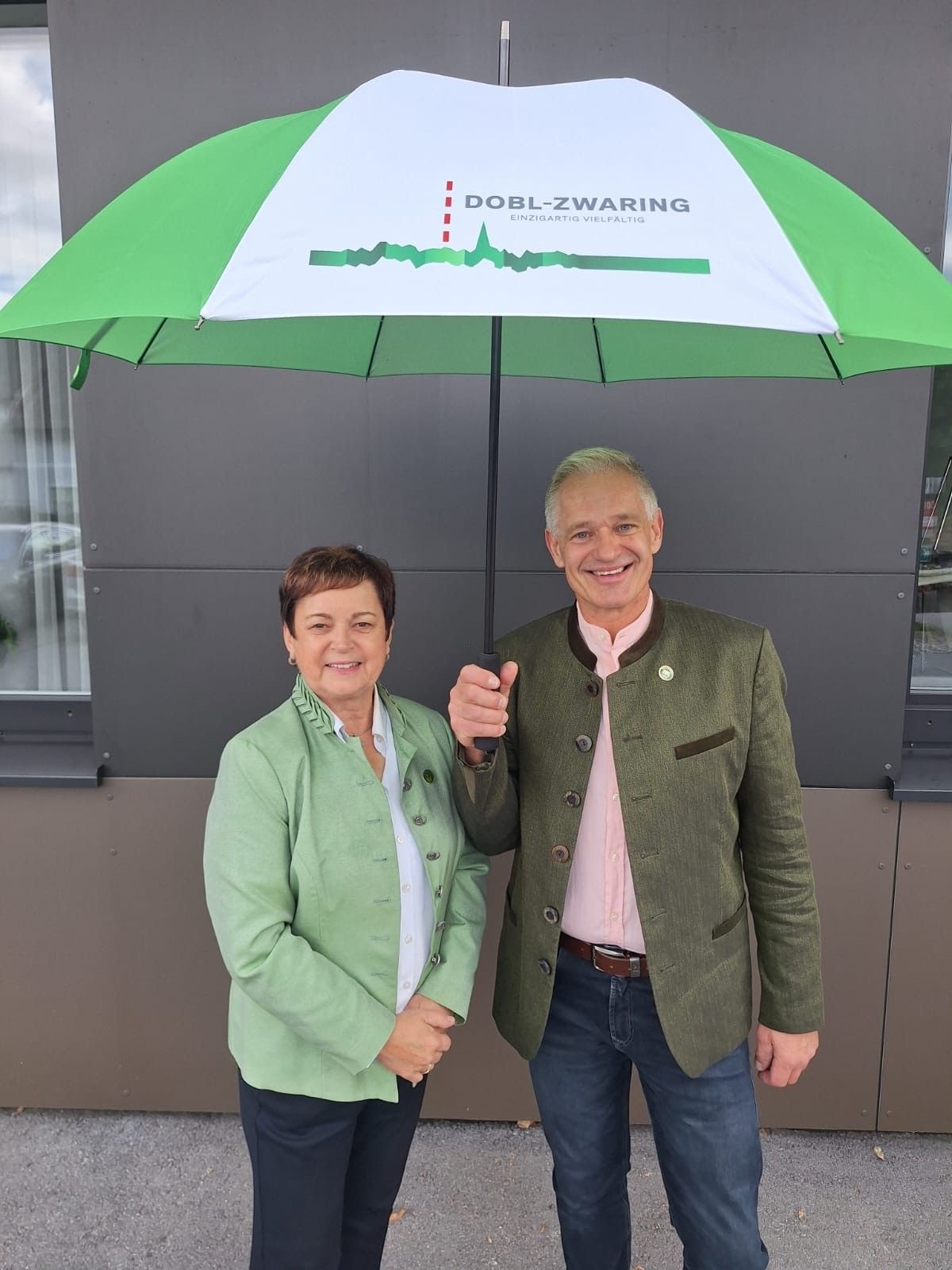 Ein Mann und eine Frau stehen nebeneinander unter einem grün-weißen Regenschirm mit dem Text Dobl-Zwaring. Sie lächeln beide und scheinen für ein Foto zu posieren.