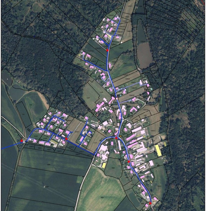 Eine Luftaufnahme eines ländlichen Gebiets mit einem netzartigen Muster von Straßen. Die Straßen sind blau und kreuzen sich mit roten Punkten, die bestimmte Punkte markieren. Gebäude sind verstreut, einige in Clustern. Die Landschaft ist überwiegend grün, mit braunen Flecken, die Felder anzeigen. Bäume umgeben das Gebiet und bilden eine natürliche Grenze.