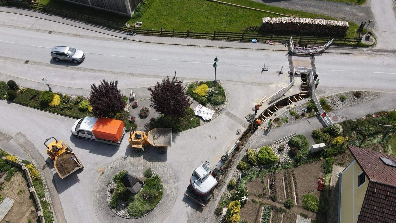 Eine Draufsicht auf eine Baustelle mit einer Straße, geparkten Maschinen, Arbeitern und einem Rasenbereich. Die Straße ist im Bau mit Barrieren und einem Holzstapel.